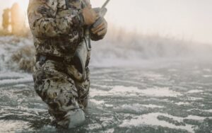 Hunter in camouflage kneeling on frozen ground holding firearm in winter landscape