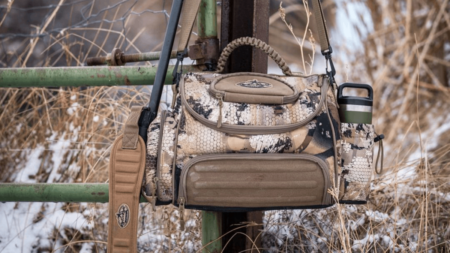 Camouflage hunting bag hanging on rusty metal fence in snowy winter landscape