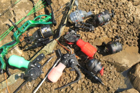 Best Bowfishing Reels of 2024, Expert Tested Bowfishing equipment including compound bow, reels, and accessories laid on rocky ground