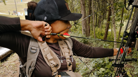 Person aiming compound bow at outdoor archery range wearing backpack and cap
