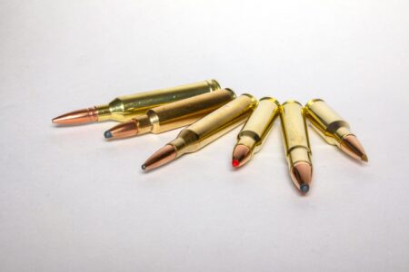 Six brass rifle cartridges with colored polymer tips arranged on white surface