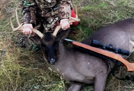 Texas Teen Harvests Ultra-Rare All-Black Whitetail Hunter in camouflage posing with harvested deer and scoped rifle outdoors.