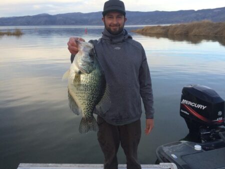 There’s a New Pending Record Black Crappie in California Man holding large crappie fish on boat with Mercury outboard motor lakeside