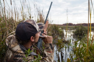 Hunter in camouflage aiming shotgun over wetland marshes during waterfowl hunting season