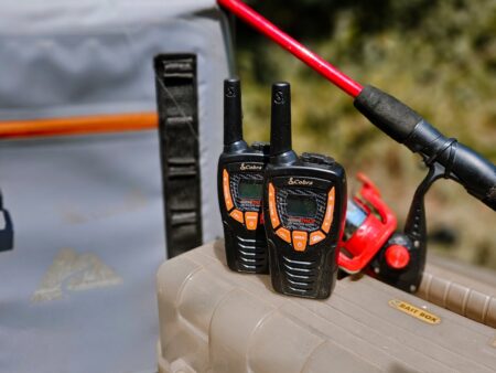 Two Cobra walkie-talkies on a bait box beside a fishing rod outdoors.