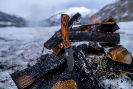 Fixed blade knife with orange black G10 handle resting on campfire logs in snow