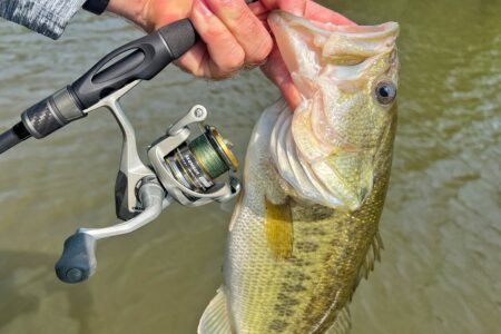 Okuma Avenger Spinning Reel, Expert Tested Angler holding largemouth bass beside Okuma Avenger spinning reel over water