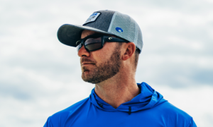 Man wearing blue hoodie, Costa sunglasses, and gray trucker hat outdoors.