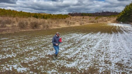 Farmer walking through muddy snowy field carrying red bag in rural countryside