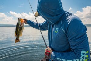 Angler in blue hoodie holds freshly caught bass over calm lake water