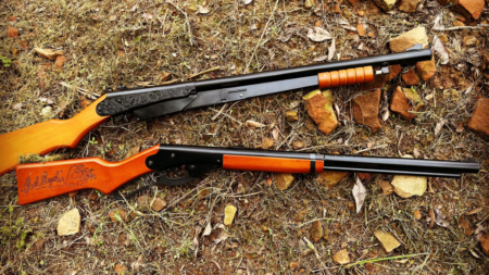 The 5 Best BB Guns for Hunting and Training Two rifles with wooden stocks lying on dry grass and rocks outdoors.