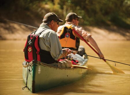 Two men fishing from a canoe on a muddy river wearing life vests.