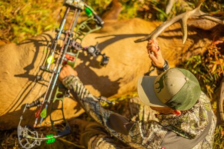 The 5 Best Bow Stabilizers of 2024 Hunter in camouflage holding compound bow beside harvested buck with antlers outdoors.