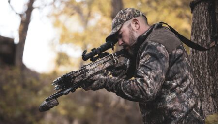Hunter in camouflage aiming crossbow with scope in autumn forest setting