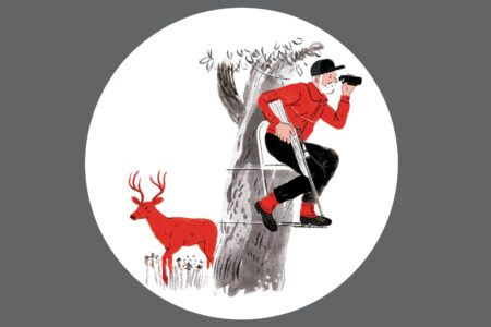 Hunter in red jacket uses binoculars in tree while red deer stands nearby.
