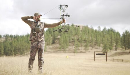 How to Shoot a Compound Bow: Archery Tips for Beginners and Beyond Hunter in camouflage aiming compound bow in open field with pine trees background