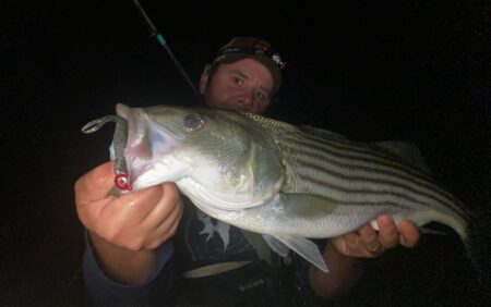 How to Sneak in Some Striped Bass Fishing on a Family Vacation This Summer Angler holding striped bass caught at night with fishing lure in mouth