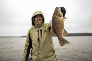 Smiling angler in rain jacket holding up a caught smallmouth bass on overcast lake