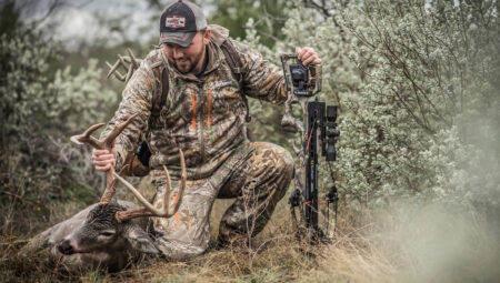Hunter in camouflage kneeling beside harvested whitetail buck holding compound bow outdoors.