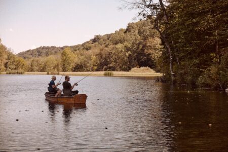 Two men fishing from a small wooden rowboat on a calm forest lake.