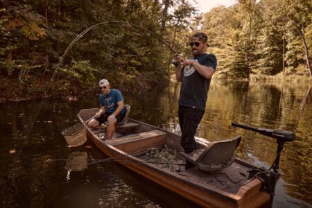 Two men fishing from a wooden boat on a calm forest river.