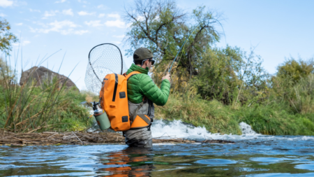 Fly fisherman wading in river wearing waders, orange backpack, and green jacket.