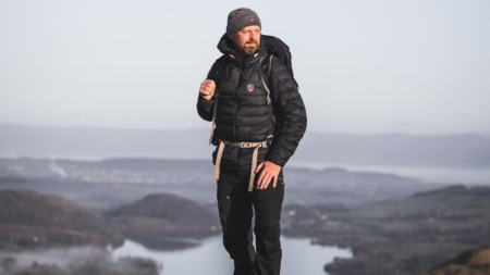 Bearded hiker in black jacket and backpack standing on misty mountain summit.