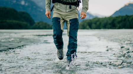 Hiker wading through shallow mountain river wearing waterproof outdoor pants and boots