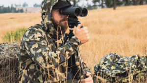 Hunter in camouflage gear using binoculars on tripod in golden grassland field