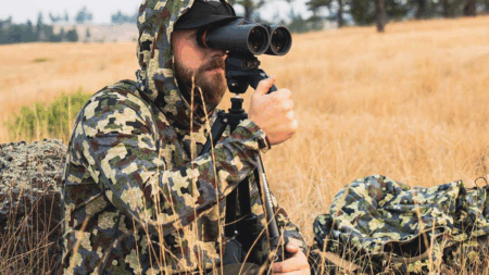Our Favorite American-Made Outdoor Gear Hunter in camouflage gear using binoculars on tripod in golden grassland field