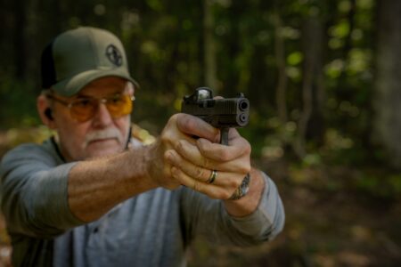 Older man aiming black pistol with red dot sight in forest setting