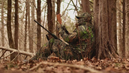 Hunter in camouflage gear sitting against tree in forest holding rifle