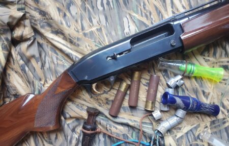 Mossy Oak camouflage hunting setup with pump-action shotgun, shotgun shells, and duck calls