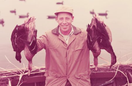 Smiling hunter holding two harvested ducks near a lake with decoys