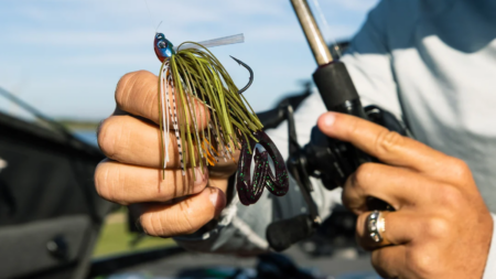 The Best Spring Bass Lures, According to Pros Angler holding colorful fishing jig lure with hooks near baitcasting reel outdoors