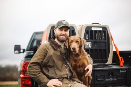 Man in hunting gear poses with Chesapeake Bay Retriever beside pickup truck outdoors.