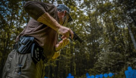 Man in tactical gear drawing pistol at outdoor shooting range in forest.