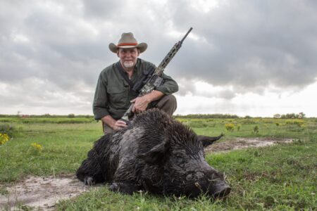 Hunter poses with harvested wild boar holding AR-style rifle in open field.