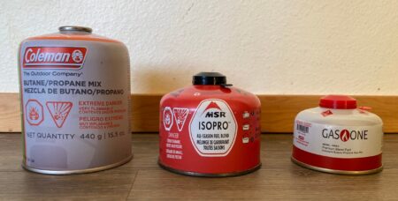 Three camping fuel canisters: Coleman, MSR IsoPro, and Gas One, on wood shelf.