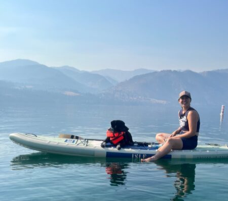 Woman and black dog in life vest sit on ISLE paddleboard on calm mountain lake.