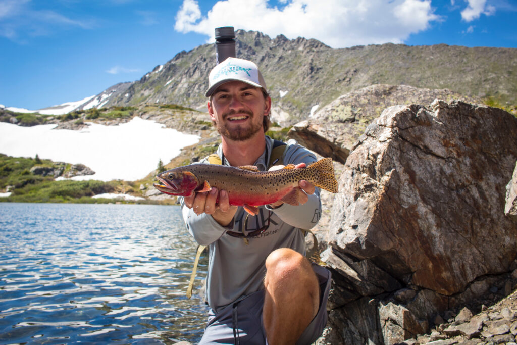 Stillwater Trout Fishing: A Beginner’s Guide Smiling man holding a colorful rainbow trout beside a mountain alpine lake