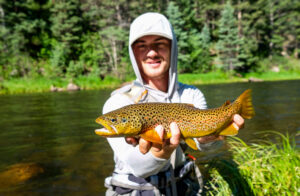 Smiling angler holding a golden brown trout beside a scenic forest river.