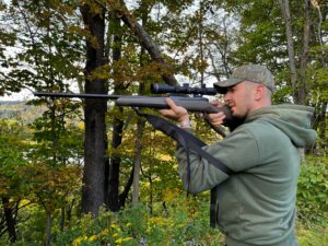 Hunter aiming scoped rifle in autumn forest wearing camouflage cap and green hoodie