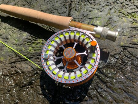 Silver and orange fly fishing reel with cork handle rod on wet rock.