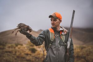 Hunter in orange cap holds harvested bird while carrying shotgun in misty hills.