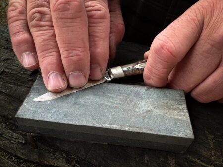 Hands sharpening a folding pocket knife on a gray whetstone outdoors
