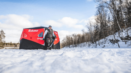 The Best Ice Fishing Shelters of 2024 Person standing beside red Eskimo ice fishing shelter on frozen snow-covered lake.