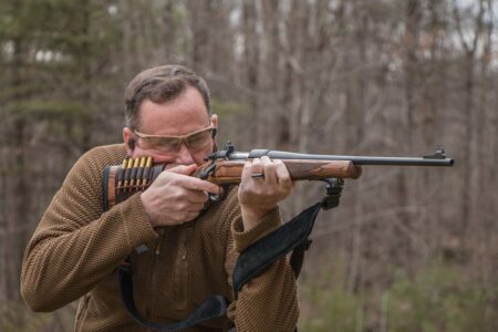 Man aiming a bolt-action rifle outdoors wearing safety glasses and ammunition bandolier.