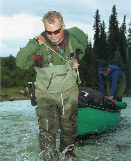 Man in waders pulling green canoe through river with companion unloading gear
