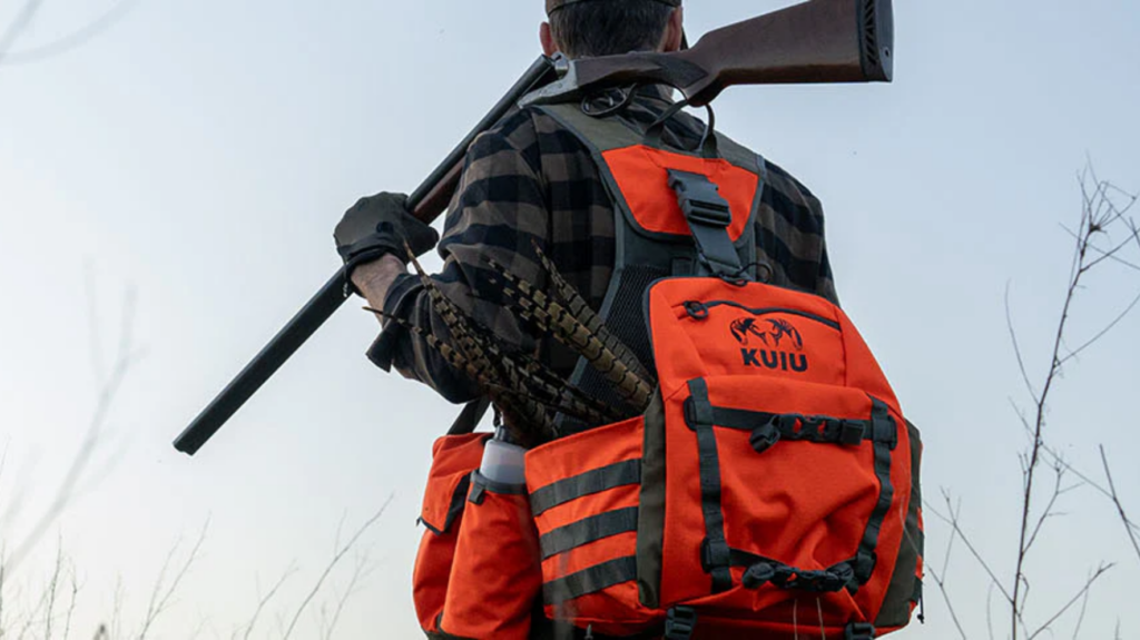 Hunter carrying rifle wearing orange KUIU backpack in outdoor hunting setting